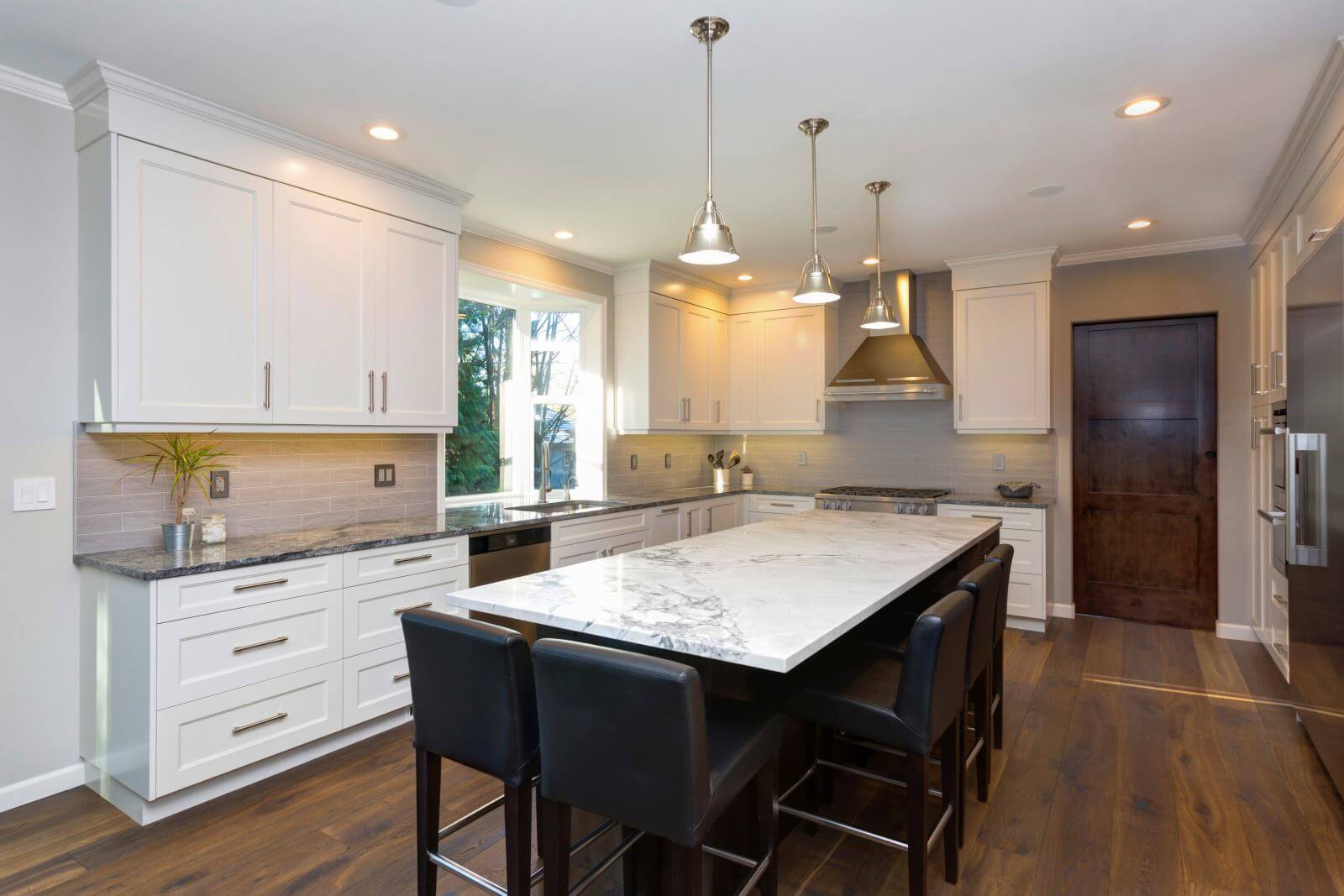 Luxury home interior boasts Beautiful black and white kitchen with custom white shaker cabinets, endless marble topped kitchen island with black leather stools over wide planked hardwood floor.
