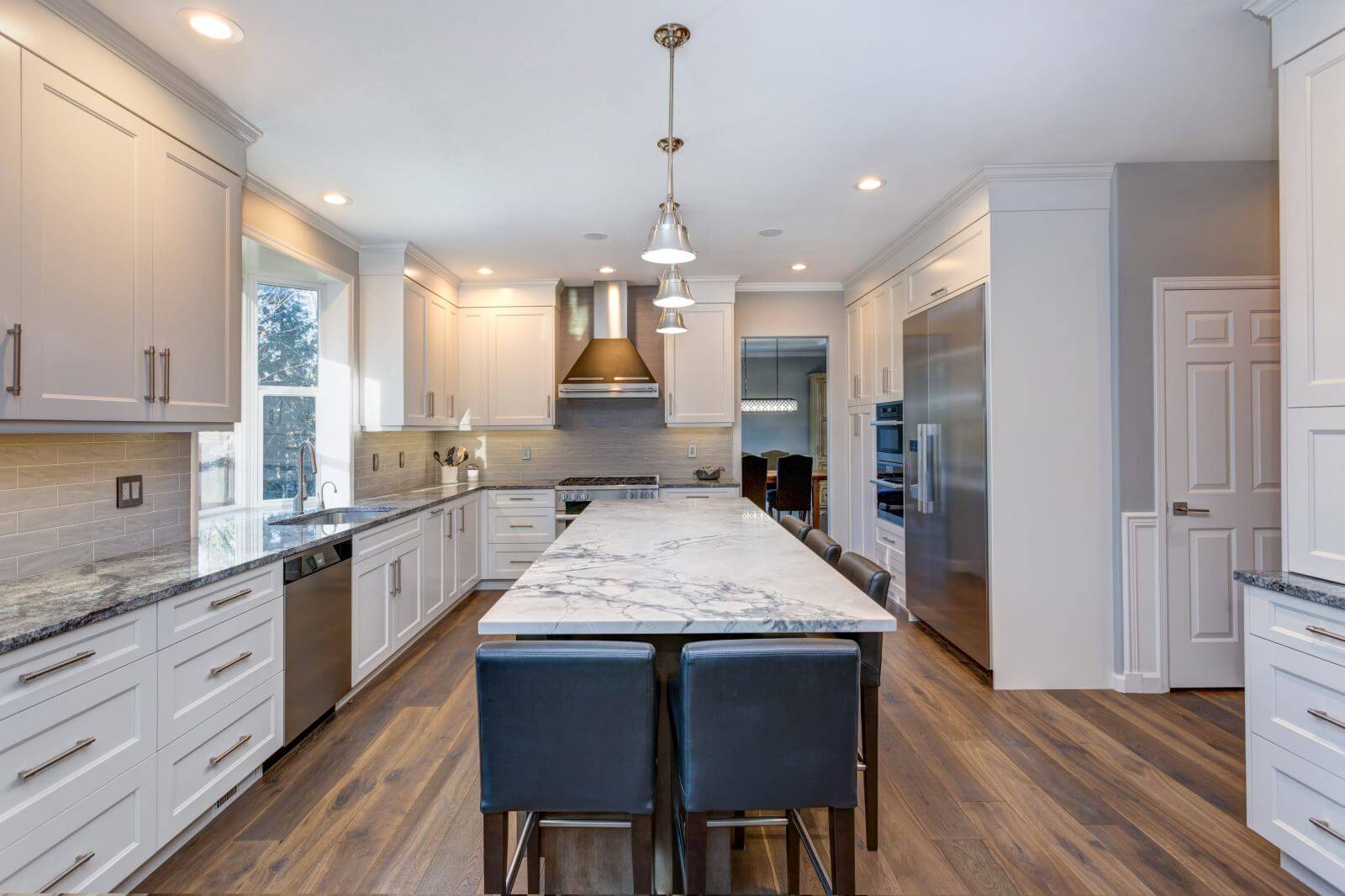 Luxury home interior boasts Beautiful black and white kitchen with custom white shaker cabinets, endless marble topped kitchen island with black leather stools over wide planked hardwood floor.