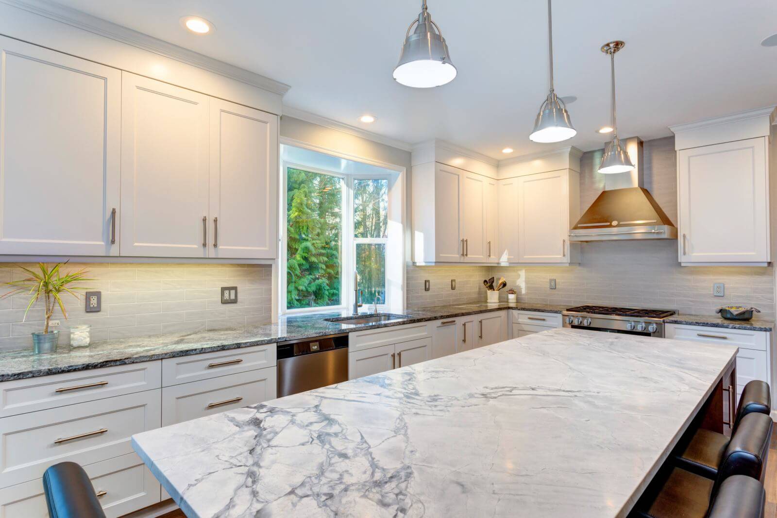 Luxury home interior boasts amazing white kitchen with custom white shaker cabinets, endless marble topped kitchen island and stainless steel appliances over wide planked hardwood floor.