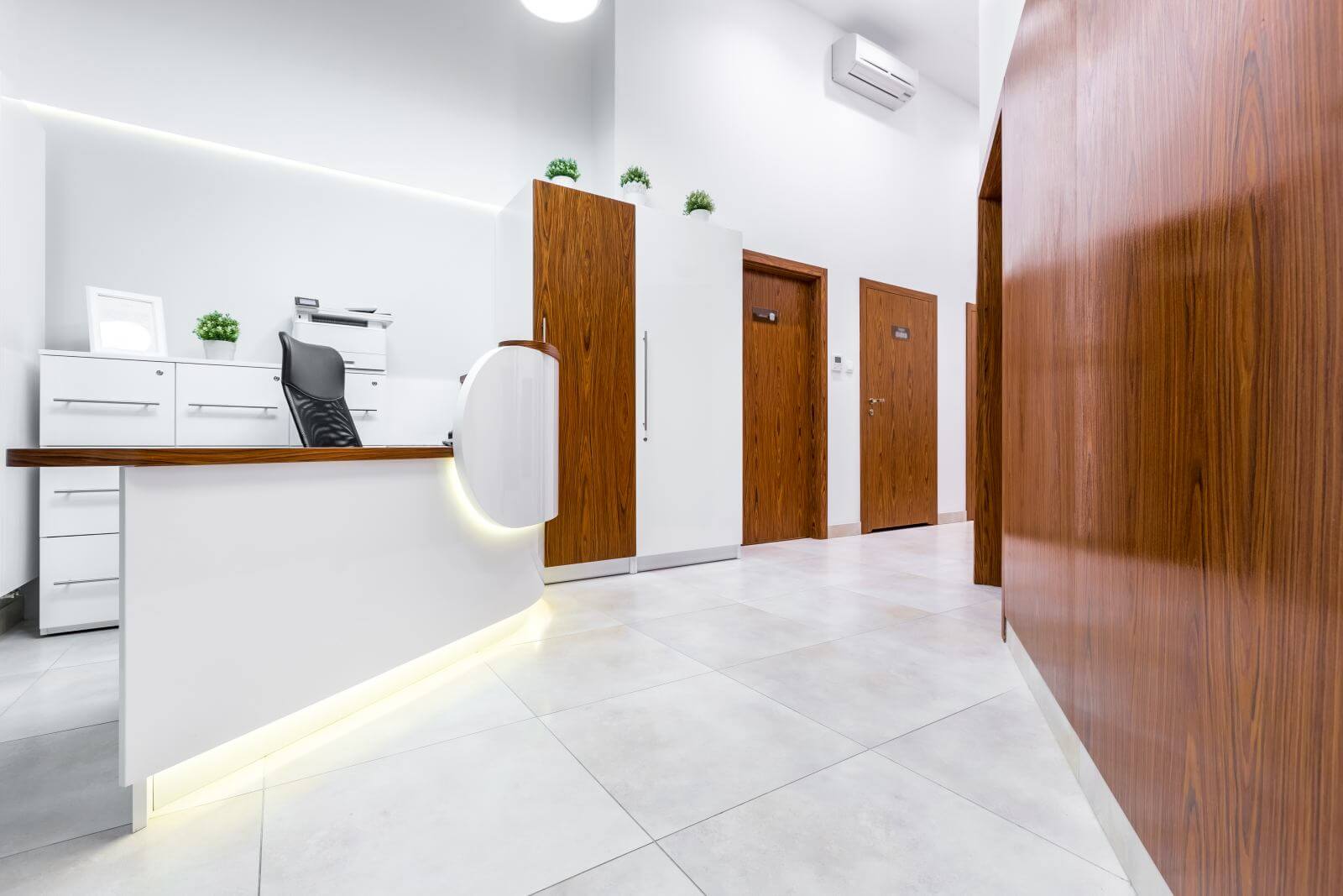 Reception of modern, private clinic with white flooring, wooden doors and desk